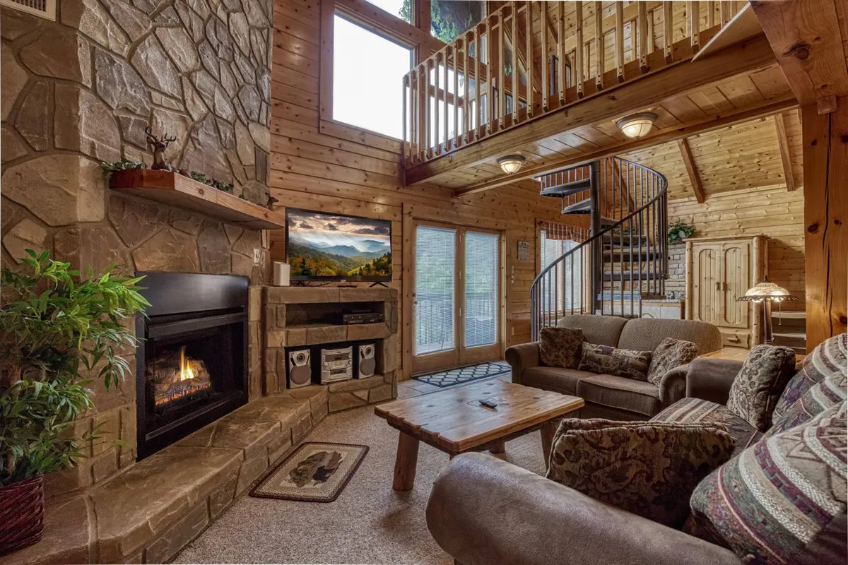 A welcoming living room featuring a prominent stone fireplace, plush seating, a spiral staircase, and expansive windows that frame stunning mountain vistas. The space glows with natural light, inviting relaxation and socializing.