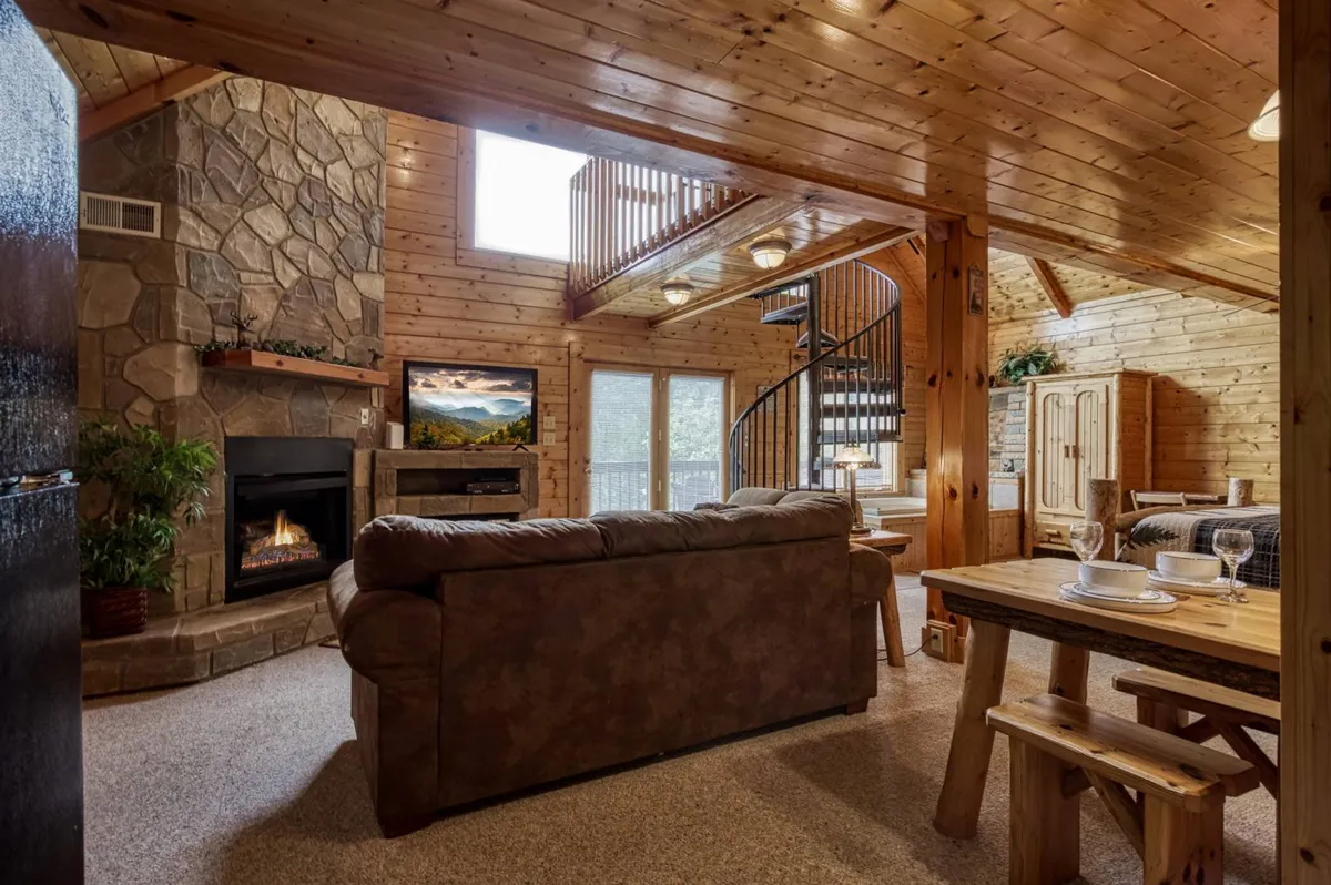 A sunlit interior highlighting a spacious dining area with a beautifully set wooden table, stone fireplace, and large windows offering sweeping scenic views of the mountains. The spiral staircase adds architectural charm, while comfortable seating creates a sense of warmth and togetherness.