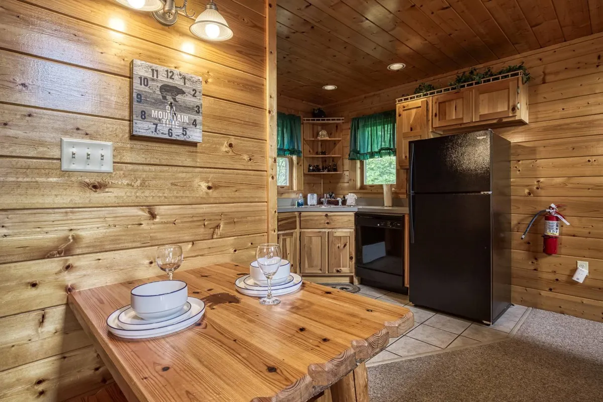 An inviting interior view featuring a wooden dining table set near the kitchen area. The space is accented with a black refrigerator, green window curtains, and warm wooden cabinets, creating a cozy and functional setting perfect for both cooking and dining. The open design connects the kitchen and dining areas, enhancing the sense of togetherness.