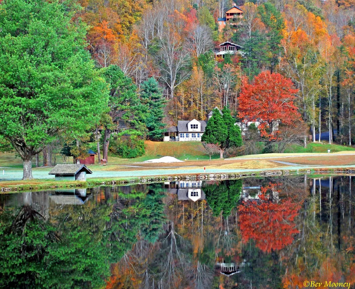 This exterior scene features a picturesque lake framed by trees and houses, with vibrant autumn foliage and a serene, glassy reflection on the water. The inviting shoreline offers ideal spots for fishing, picnicking, or simply enjoying panoramic mountain and lake views. The overall atmosphere is calm and welcoming, perfect for family exploration and outdoor fun.