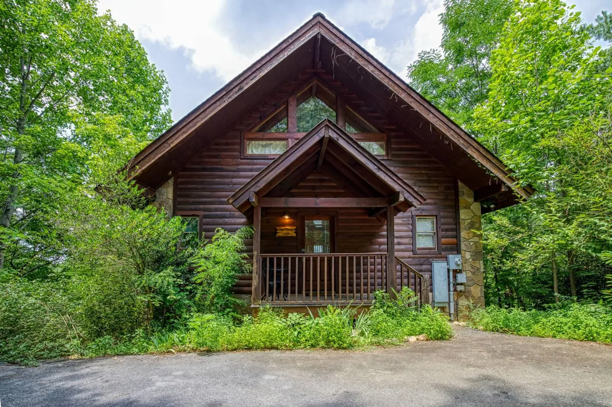 The image features a charming log cabin perched atop a mountain, with a welcoming front porch surrounded by lush natural landscaping. The setting highlights the inviting porch as a perfect spot for relaxing, enjoying sweeping views, and basking in serene mountain ambiance. The surrounding woods and open sky suggest opportunities for stargazing, sunset watching, and connecting with nature.