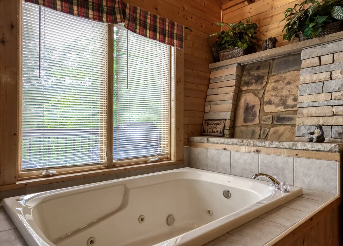 A serene bathroom scene with a spacious jacuzzi tub nestled against a natural stone feature wall and flanked by potted plants. A window with blinds lets in gentle natural light, enhancing the tranquil, spa-like setting.