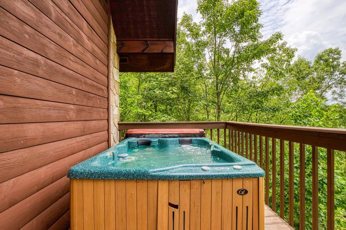This exterior image showcases a hot tub on a wooden deck, surrounded by towering trees. The deck offers an inviting space for families to unwind, soak in warmth, and immerse themselves in nature, while hints of sunset light filter through the trees. The tranquil outdoor setting suggests both relaxation and the opportunity for social connection in a scenic environment.