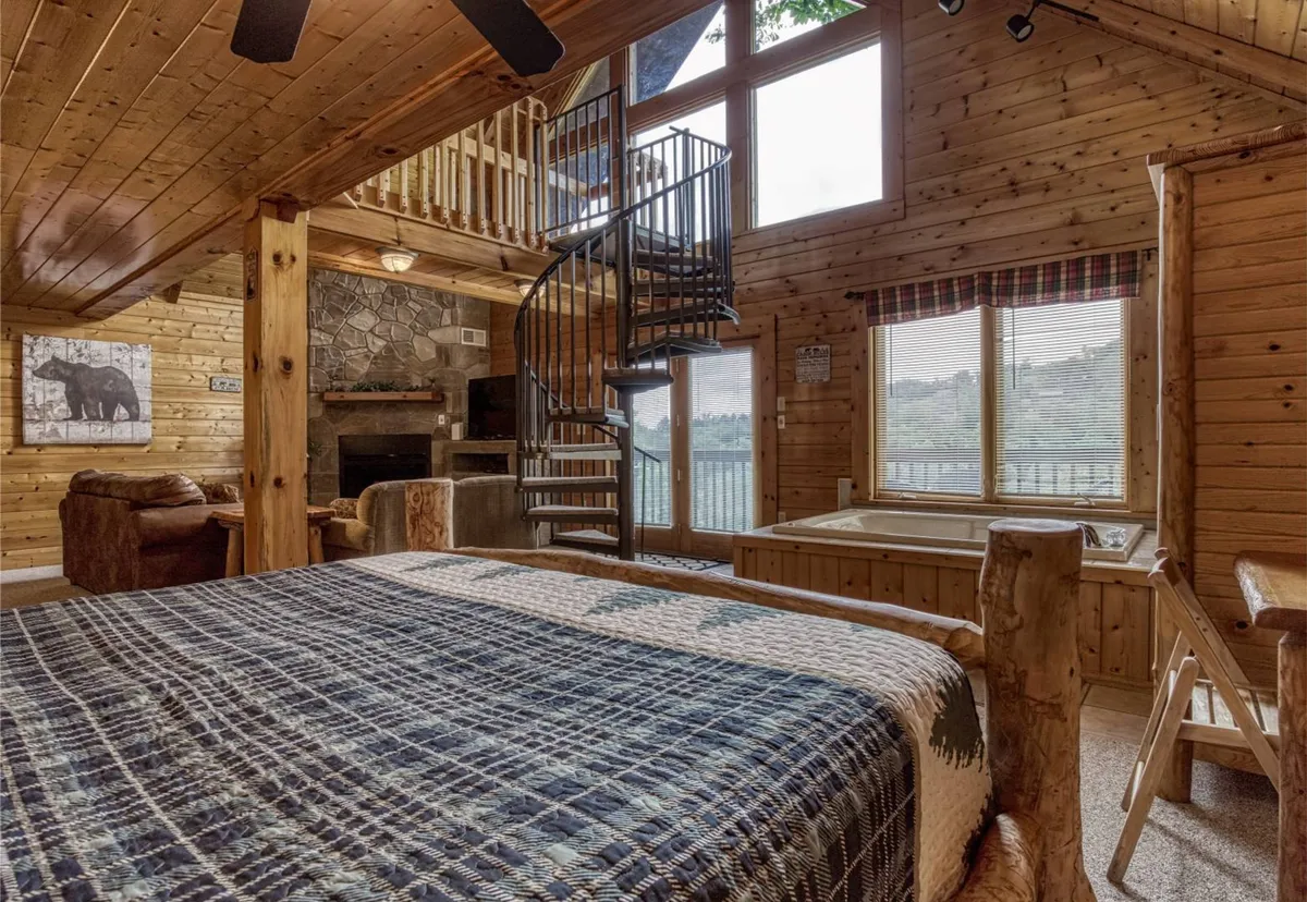 A charming interior with a spiral staircase, large windows offering scenic views, a warmly dressed bed with a quilted spread, and a stone fireplace. The inviting setup suggests cozy mornings and evenings spent in comfort and seclusion.