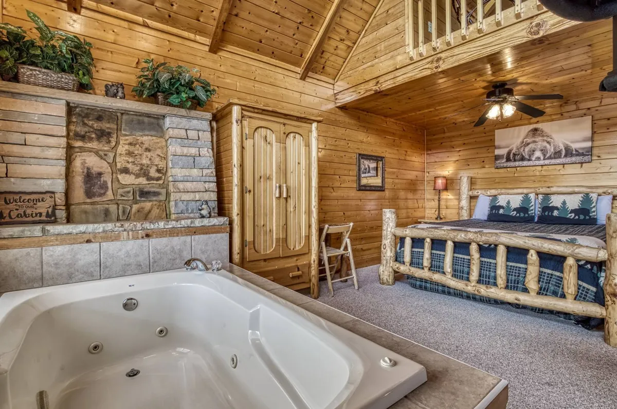 A warm, welcoming interior featuring a rustic log bed beside a stone fireplace, with a luxurious jacuzzi tub positioned nearby. Wooden accents and a ceiling fan add to the cozy ambiance, while soft lighting and plush bedding suggest relaxation and comfort.